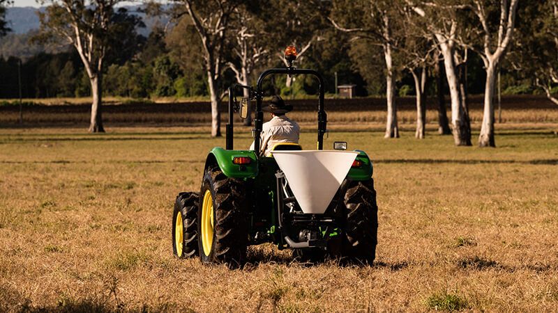 Male farmer operating a Rapid Spray FertLink spreader attached to a John Deere tractor, applying fertiliser across a field